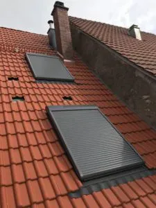 Zwei geschlossene Rollläden an Fenstern in einem schrägen roten Ziegeldach zeugen von der handwerklichen Geschicklichkeit der Dachdeckerei.