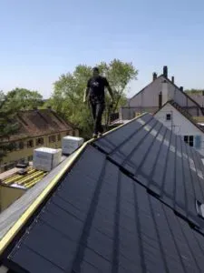 Eine Person aus einer Dachdeckerei läuft auf dem First eines mit Solarmodulen bestückten Schrägdachs, im Hintergrund umgeben von Häusern und Bäumen.