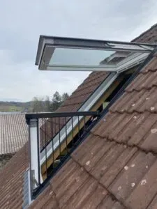 Ein von einer Dachdeckerei fachmännisch eingebautes Dachfenster öffnet sich nach außen und bildet einen kleinen Balkon auf einem schrägen Ziegeldach.