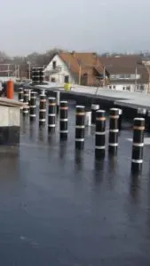 Mehrere schwarz-weiße Poller stehen auf einem nassen, flachen Dach, wahrscheinlich Teil einer Dachterrasse, im Hintergrund sind Häuser zu sehen.