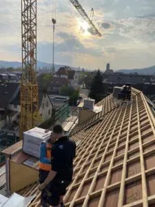 Zwei Dachdecker auf einem Dach im Bau, dahinter Kran und Stadt im Abendlicht