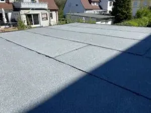 Flachdach mit grauer Dacheindeckung, fachmännisch von einer erfahrenen Dachdeckerei errichtet, umgeben von Häusern und Bäumen unter klarem Tageslicht.