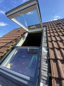 Ein von einer Dachdeckerei fachmännisch eingebautes Dachfenster auf einem Schrägdach steht unter einem strahlend blauen Himmel offen.