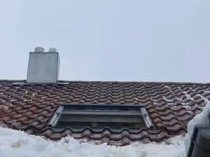 Schneebedecktes Ziegeldach eines fachmännischen Dachdeckers mit geschlossenem Fenster und Schornstein unter bewölktem Himmel.