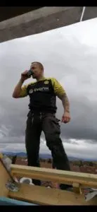 Mann steht im Freien auf Holzbalken, möglicherweise auf einem Dachdeckereigelände, und trinkt unter bewölktem Himmel aus einer Flasche.