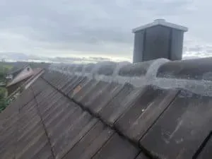 Ziegeldach mit ausgebessertem Mörtel am First, im Hintergrund ein Schornstein