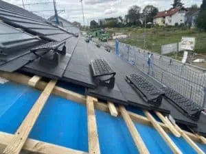 Ein teilweise errichtetes Dach mit schwarzen Ziegeln, Holzbalken und darauf installierten Dachstufen zeigt handwerkliche Dachdeckerei in Aktion.