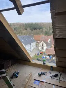 Blick von einem Dachboden, der gerade von einem erfahrenen Dachdecker ausgebaut wird, auf Dächer und ein Waldgebiet in der Ferne.