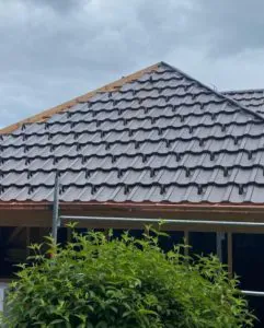 Haus mit grauem Ziegeldach unter bewölktem Himmel, vorne grüne Büsche