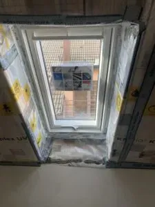 Ein von der Fachfirma Dachdeckerei schräg eingebautes Dachfenster, umgeben von Dämmmaterial und Rohbauwänden.