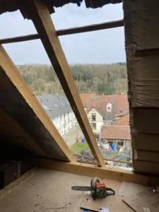 Blick durch ein geöffnetes Dach mit Holzbalken auf Häuser, daneben eine Kettensäge