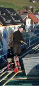 Dachdecker mit rotem Elektrowerkzeug und Metallplatten auf einem Dach, dahinter Häuser