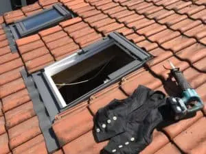Offenes Fenster auf einem Ziegeldach, mit Werkzeug und einer Arbeitsweste in der Nähe, bereit für Wartungsarbeiten an der Dachdeckerei.