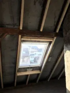 Dachfenster in einem Schrägdach mit freiliegenden Holzbalken, in fachmännischer Dachdeckerei gefertigt, mit Blick auf die Häuser draußen.