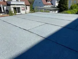 Flachdach mit überlappenden grauen Platten, dahinter Häuser und Bäume