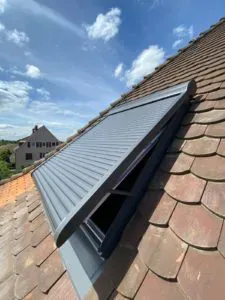 Dachfenster mit teilweise geöffnetem Metallrollladen auf einem Ziegeldach