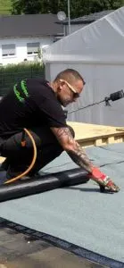 Dachdecker mit Handschuhen kniet auf einem Flachdach und verlegt Dachbahnen