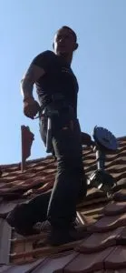 Ein Mensch in Arbeitskleidung steht mit Werkzeug auf einem Ziegeldach und blickt über die Schulter – eine typische Szene in der Welt der Dachdeckerei.