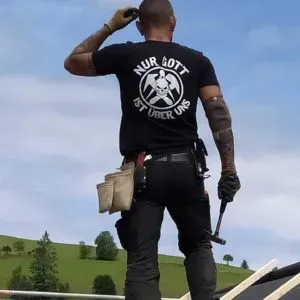 Ein Arbeiter steht auf einem Dach und stellt eine Dachdeckerei dar; Auf seinem T-Shirt steht „Nur Gott ist über uns“ mit einer Totenkopfgrafik.
