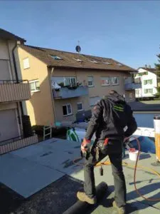 Ein Arbeiter in Sicherheitsausrüstung steht an einem sonnigen Tag auf einem Dach und benutzt Werkzeuge während eines Dachdeckerei-Projekts.