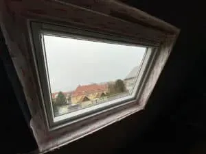 Ein schräges Dachfenster, das mit fachmännischer Dachdeckerei gefertigt wurde, rahmt den Blick auf Häuser und Bäume unter einem bewölkten Himmel.