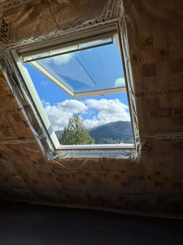 Ein offenes Dachfenster in einem unausgebauten Dachgeschoss bietet einen malerischen Blick auf Berge, Bäume und einen blauen Himmel mit Wolken.