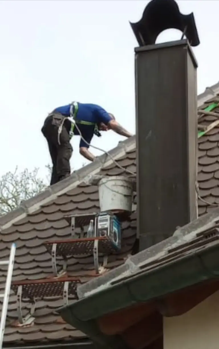 Ein Arbeiter in Schutzkleidung repariert ein Ziegeldach neben einem Schornstein. In der Nähe stehen Werkzeuge und ein Eimer.