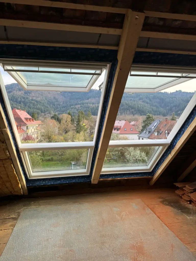 Blick durch offene Dachfenster auf Dächer, Bäume und Berge in der Ferne unter einem bewölkten Himmel.