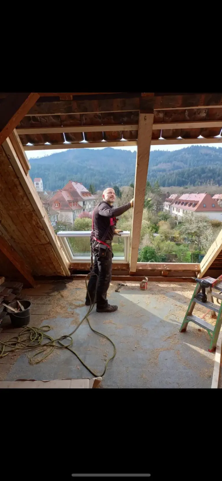 Ein Mann arbeitet an einem hölzernen Dachstuhl, im Hintergrund eine malerische Stadt und bewaldete Hügel.