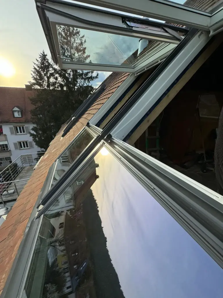 Geöffnete Dachfenster reflektieren den Himmel bei Sonnenuntergang auf einem schrägen Dach und bieten Ausblick auf die benachbarten Häuser und Bäume.