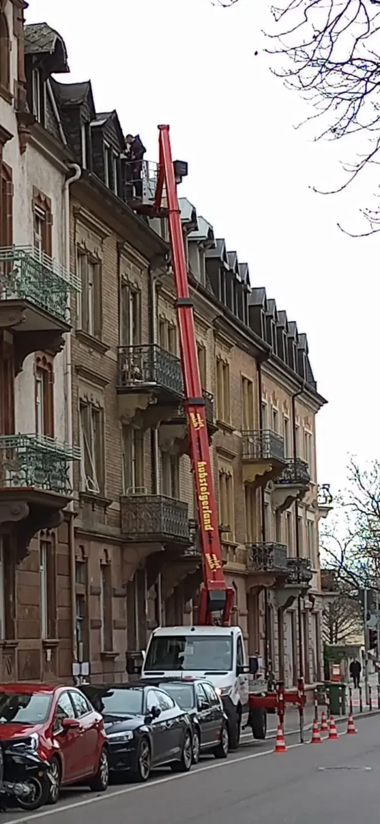 Ein roter Kran hebt eine Person auf das Dach eines braunen Wohnhauses in einer Stadtstraße.