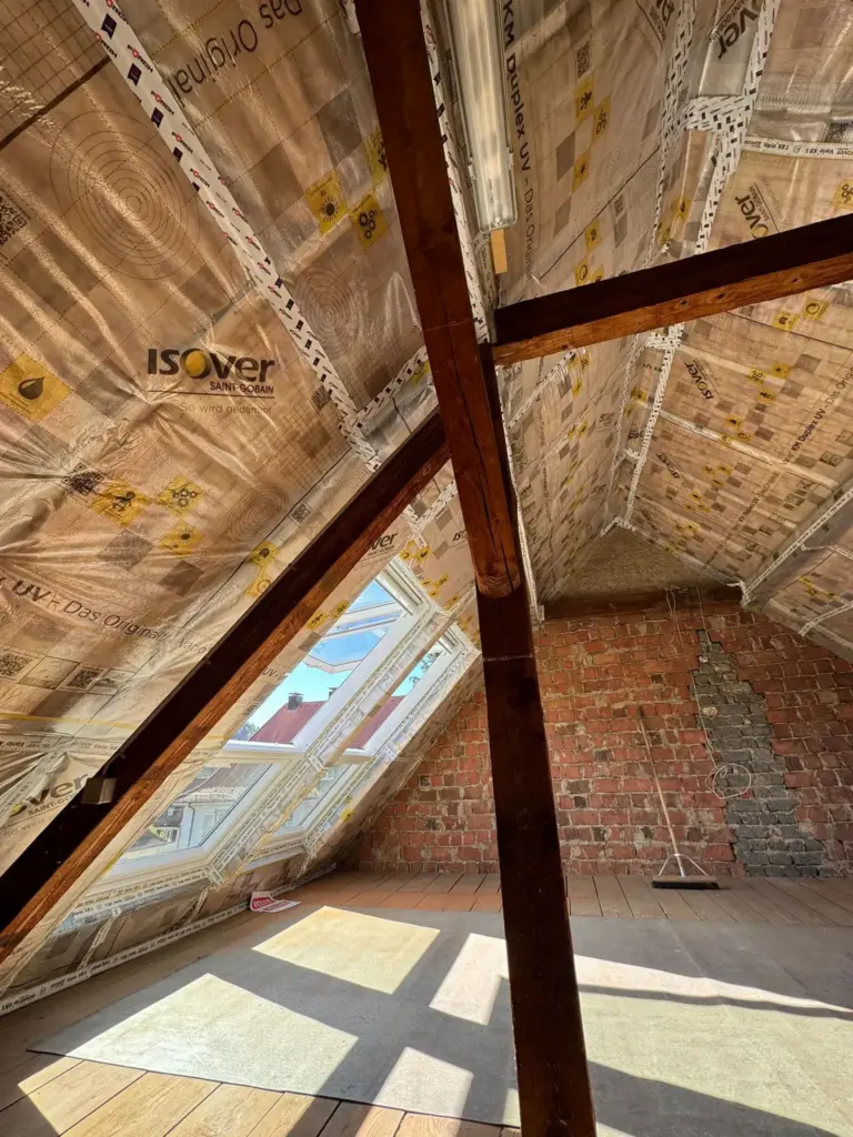 Dachboden mit schrägen Balken, freiliegenden Ziegelwänden und Dämmung an der Decke; Sonnenlicht fällt durch das Oberlicht ein.
