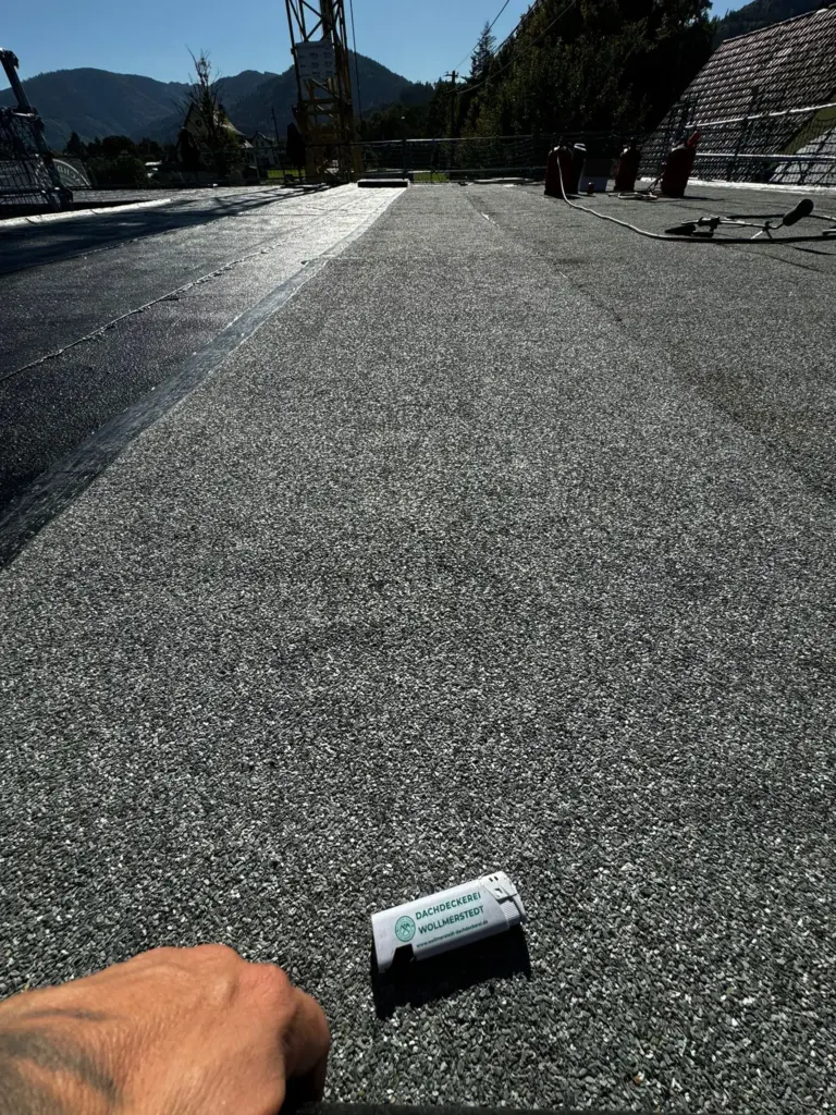 Eine Hand hält Dachmaterial auf einem flachen Kiesdach mit Bergen und Baumaschinen im Hintergrund.