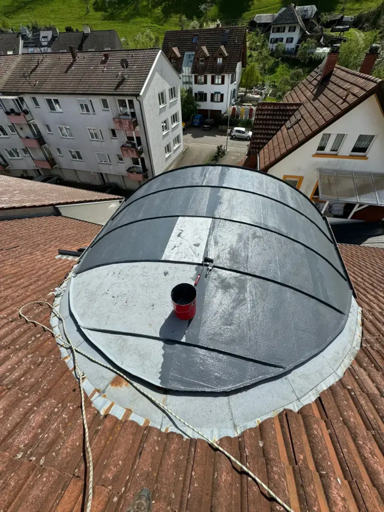 Ein gewölbtes Dach wird mit einer schwarzen wasserfesten Beschichtung gestrichen; oben sind eine Farbrolle und ein Eimer zu sehen.