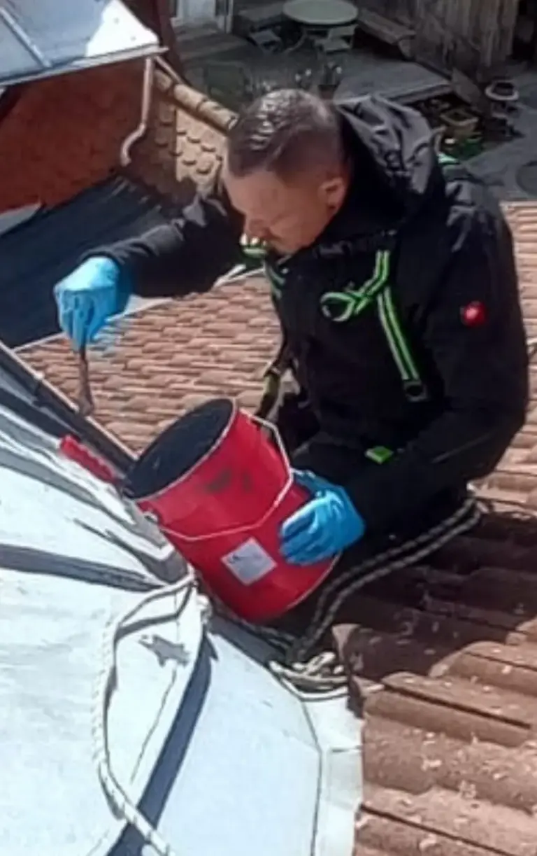 Eine Person mit blauen Handschuhen streicht mit einem Pinsel ein Dach, hält einen roten Farbeimer in der Hand und sitzt auf Ziegeln.