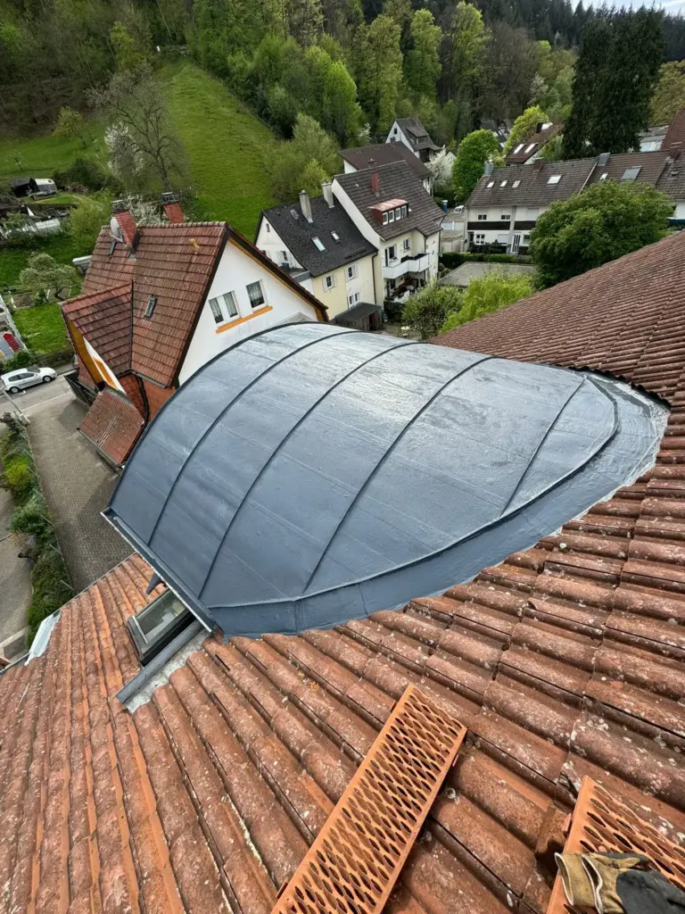 Auf einem roten Ziegeldach in einem Wohngebiet wird ein geschwungenes graues Dachteil montiert.