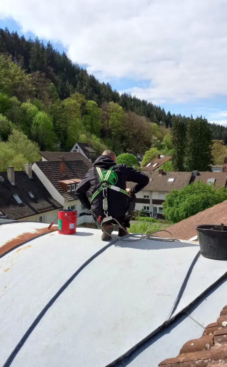 Ein Arbeiter in Sicherheitsausrüstung kniet auf einem Dach, umgeben von Bäumen und Häusern in einer hügeligen Gegend.